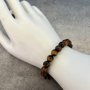 May include: A brown and gold tiger's eye beaded bracelet. The bracelet is made of round beads and is worn on a white mannequin hand.