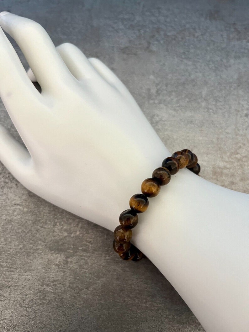 May include: A brown and gold tiger's eye beaded bracelet. The bracelet is made of round beads and is worn on a white mannequin hand.