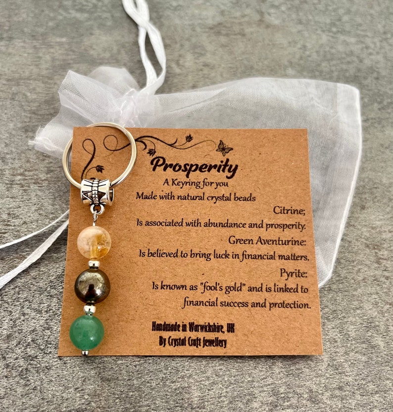 Pode incluir: Um cart&atilde;o castanho com o texto "Prosperity" e uma descri&ccedil;&atilde;o dos cristais usados no chaveiro. O chaveiro apresenta um amuleto de prata com tr&ecirc;s contas: uma citrina laranja claro, uma pirita castanho escuro e uma aventurina verde. O chaveiro est&aacute; sobre um fundo branco.