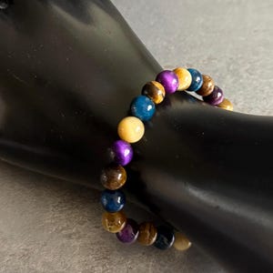Puede incluir: Un brazalete de cuentas con piedras preciosas de colores. El brazalete incluye cuentas en tonos azules, morados, amarillos y marrones. El brazalete se muestra sobre una superficie negra. Las cuentas miden aproximadamente 1 cm de di&aacute;metro.