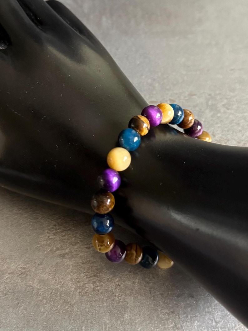 Puede incluir: Un brazalete de cuentas con piedras preciosas de colores. El brazalete incluye cuentas en tonos azules, morados, amarillos y marrones. El brazalete se muestra sobre una superficie negra. Las cuentas miden aproximadamente 1 cm de di&aacute;metro.