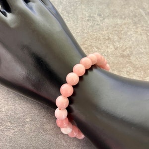 Op de afbeelding: Een lichtroze kralenarmband. De armband is gemaakt van ronde, gepolijste kralen in verschillende roze tinten. De armband wordt getoond op een zwarte mannequinhand tegen een grijze achtergrond.