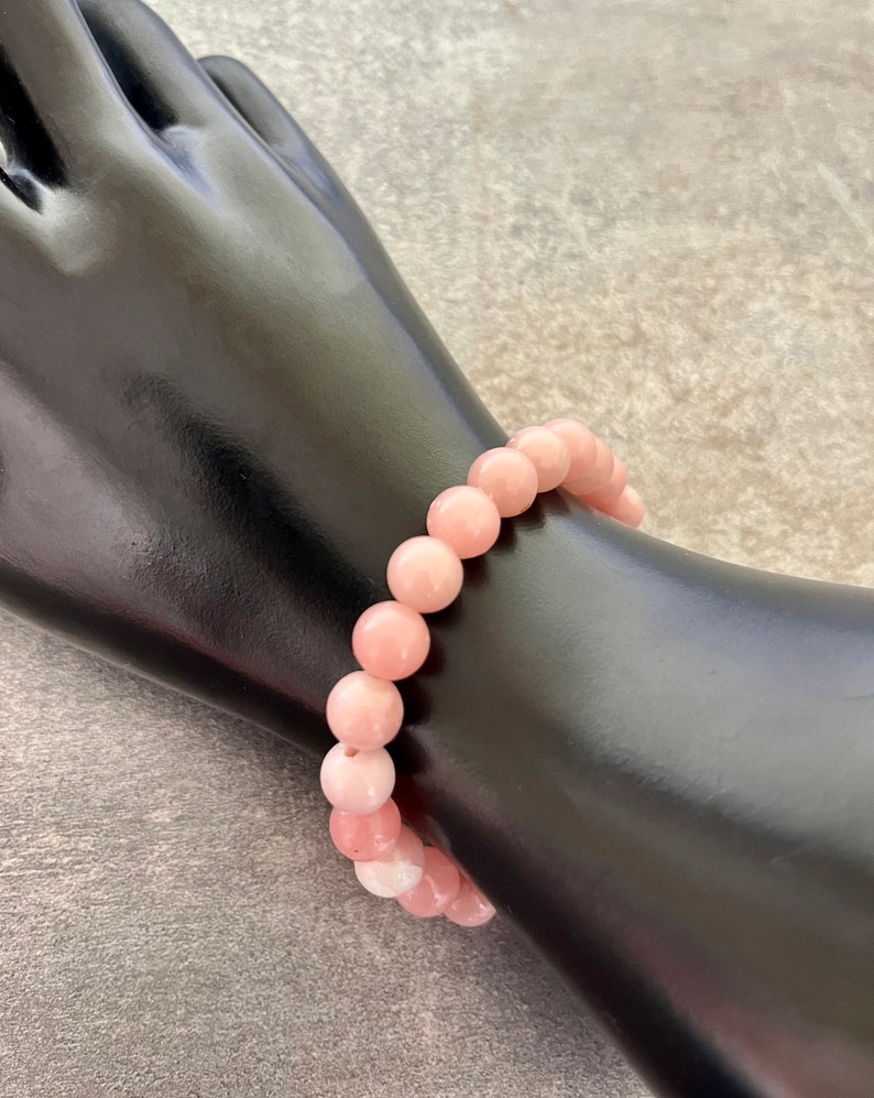 Op de afbeelding: Een lichtroze kralenarmband. De armband is gemaakt van ronde, gepolijste kralen in verschillende roze tinten. De armband wordt getoond op een zwarte mannequinhand tegen een grijze achtergrond.