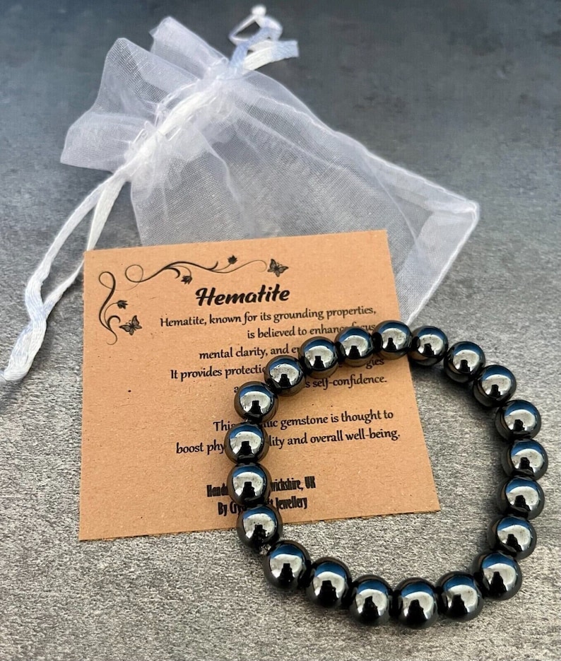 May include: Hematite bracelet made of black beads. The bracelet is on a brown card with the text "Hematite" and information about the stone's properties. The card is on a white background.