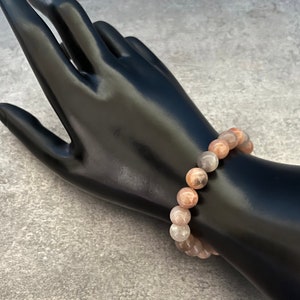 May include: A pink, gray, and white gemstone bracelet with round beads. The bracelet is on a black mannequin hand.