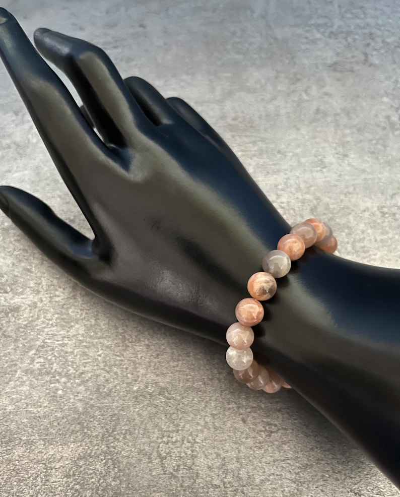 May include: A pink, gray, and white gemstone bracelet with round beads. The bracelet is on a black mannequin hand.