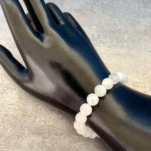 May include: A white beaded bracelet with round, opaque beads. The bracelet is displayed on a black mannequin hand against a gray background. The beads appear to be approximately 0.5 inches in diameter.