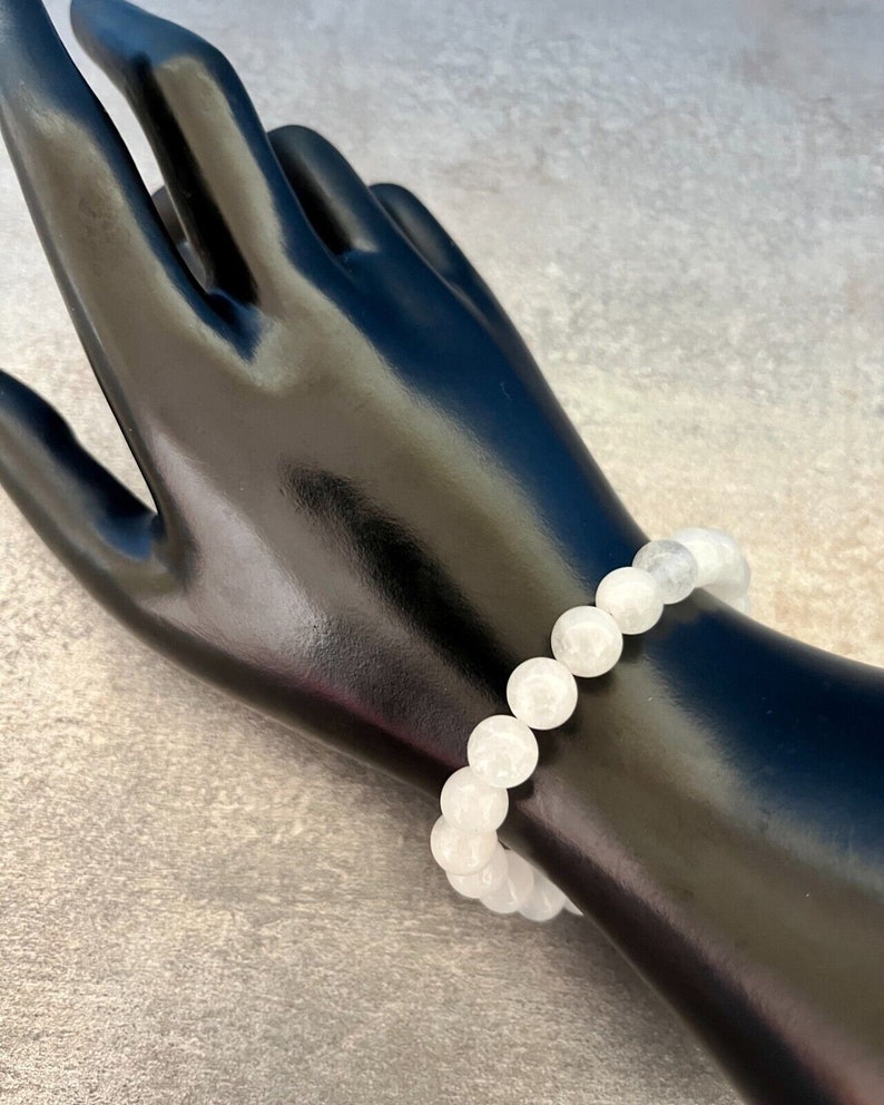 May include: A white beaded bracelet with round, opaque beads. The bracelet is displayed on a black mannequin hand against a gray background. The beads appear to be approximately 0.5 inches in diameter.