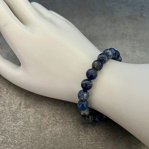 May include: A blue and white lapis lazuli beaded bracelet. The bracelet is made of round beads and is worn on a white mannequin hand.