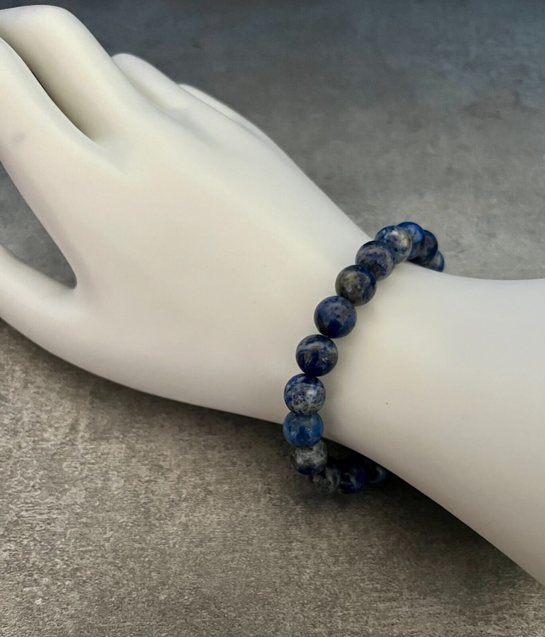 May include: A blue and white lapis lazuli beaded bracelet. The bracelet is made of round beads and is worn on a white mannequin hand.
