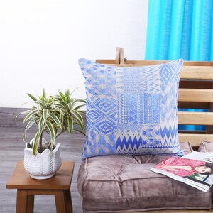 May include: A square cushion with a blue and beige geometric design. The cushion is placed on a mauve seat, beside a small wooden table with a potted plant. A magazine rests on the seat.