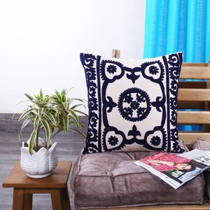 Peut inclure: Coussin décoratif crème avec un motif floral brodé bleu marine. Le coussin carré repose sur un coussin mauve. Une petite plante en pot et un magazine sont également visibles.