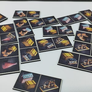 Puede incluir: Fichas de juego con ilustraciones de cajas, edificios y maquinaria, dispersas sobre una superficie blanca. Las fichas rectangulares tienen un borde negro. En el fondo se ve un tablero de juego, lo que sugiere un juego de mesa.