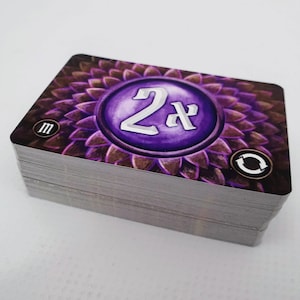May include: A stack of purple and brown game cards with a circular design featuring the text "2x" in silver lettering. The card also has a small "M" and a circular arrow symbol.