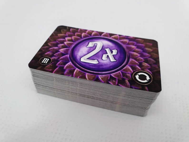 May include: A stack of purple and brown game cards with a circular design featuring the text "2x" in silver lettering. The card also has a small "M" and a circular arrow symbol.