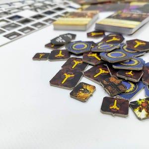 May include: A collection of board game components, including wooden tokens and card stacks. The tokens feature various designs with yellow and blue accents. The game pieces are scattered on a white surface, suggesting gameplay.