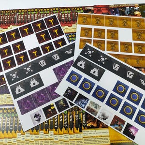May include: A collection of game components, including cards and sticker sheets. The sticker sheets feature various symbols, including skulls, flames, and circular designs with blue and gold accents. The cards have text and graphics in a variety of colors.