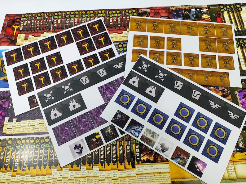 May include: A collection of game components, including cards and sticker sheets. The sticker sheets feature various symbols, including skulls, flames, and circular designs with blue and gold accents. The cards have text and graphics in a variety of colors.