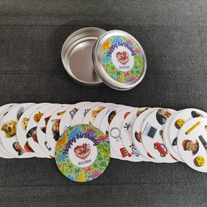May include: A set of round cards with various images and a tin container. The cards feature images of animals, people, and objects. The container lid and one card display the text "Happy birthday" with a date. The cards are arranged on a dark gray surface.