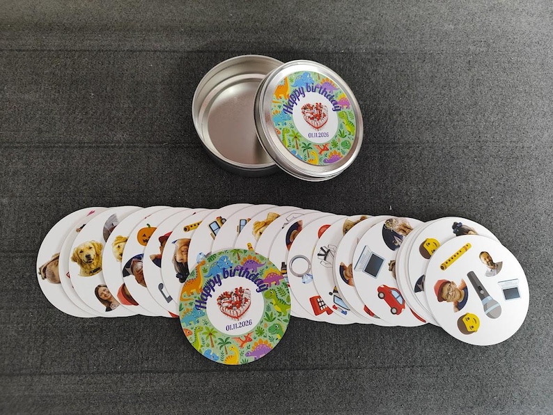 May include: A set of round cards with various images and a tin container. The cards feature images of animals, people, and objects. The container lid and one card display the text "Happy birthday" with a date. The cards are arranged on a dark gray surface.