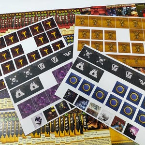 May include: A collection of printed game components, including cards and sticker sheets. The cards feature various symbols and illustrations in colors like yellow, purple, and blue. The sticker sheets have icons and images for game play.