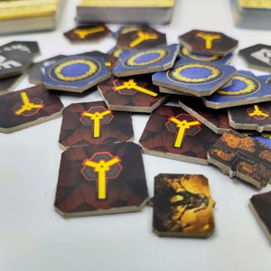 May include: A collection of game tokens with various designs. Some tokens are square with a yellow symbol on a maroon background. Others are hexagonal with a blue and gold design. A few have a dragon illustration.