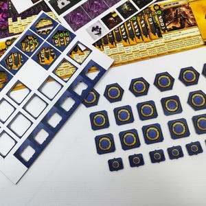 May include: A collection of board game components, including die-cut cardboard pieces and game cards. The pieces are primarily blue with gold and yellow accents. Various shapes are present, including hexagons, squares, and circles.
