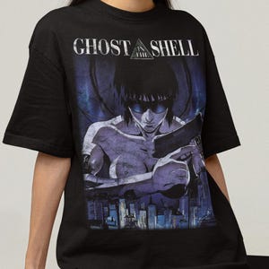 Puede incluir: Camiseta negra con un gráfico de la serie de anime Ghost in the Shell. El diseño incluye un personaje sosteniendo un arma, con el texto "GHOST SHELL" arriba. La camiseta se usa con pantalones cortos grises.