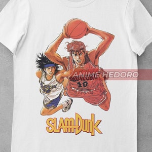 May include: White t-shirt featuring a graphic print of two basketball players from the anime "Slam Dunk". The players are in mid-air, one is shooting a basketball and the other is defending. The text "Slam Dunk" is printed in yellow below the players.