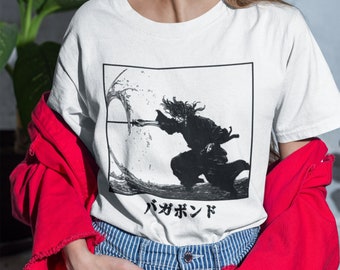 Op de afbeelding: Wit T-shirt met een zwart-witte illustratie van een persoon die een zwaard vasthoudt. De illustratie is omgeven door een zwarte rand. De tekst "バガボンド" is onder de illustratie gedrukt.