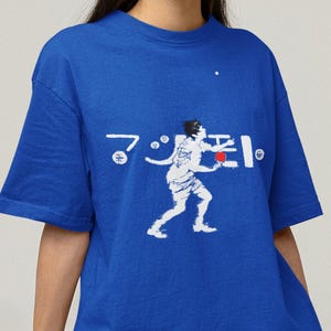 Pode incluir: Camiseta azul royal com um gráfico branco de uma pessoa praticando um esporte e texto japonês. A pessoa está segurando uma bola vermelha. A camiseta é combinada com shorts cinza.
