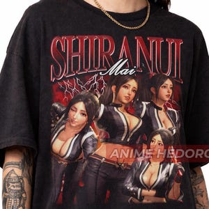 Könnte beinhalten: Dunkelgraues T-Shirt mit einer Grafik einer Frau und dem Text "SHIRANUI Mai" in Rot und Weiß. Das Design beinhaltet Blitze und die Worte "ANIME HEDORO".