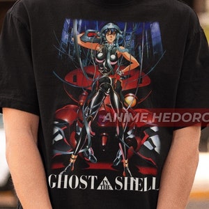 Può includere: Maglietta nera con una stampa grafica di una donna in un abito nero e rosso in piedi davanti a un grande robot. Il testo "Ghost in the Shell" è stampato sotto la grafica.