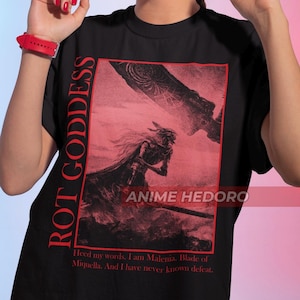 May include: Black t-shirt with a red graphic print of a character from the video game Elden Ring. The graphic includes the text "ROT GODDESS" and "ANIME HEDORO". The text below the graphic reads "Heed my words. I am Malenia. Blade of Miquella. And I have never known defeat."