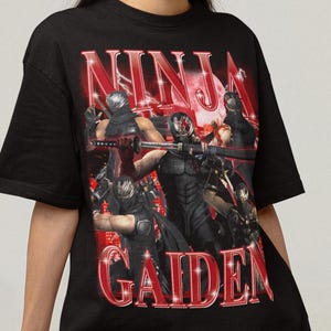 May include: Black t-shirt featuring a graphic print of ninjas from the video game Ninja Gaiden. The graphic is in red and white with a glittery effect. The text "NINJA GAIDEN" is printed in large, bold letters.