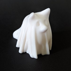 May include: A small, white ceramic ghost figurine with a draped sheet design. The ghost has a simple, rounded form with a slightly raised head and a small, rounded nose. The figurine is set against a dark background.