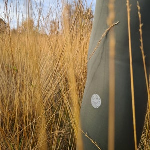 Könnte beinhalten: Nahaufnahme einer grünen, wasserdichten Hose mit weißem Logo. Die Hose ist teilweise von hohem, trockenem Gras verdeckt.