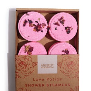 May include: A cardboard box containing four pink shower steamers, each decorated with dried rose petals. The box is printed with the words "Ancient Wisdom" and "Love Potion Shower Steamers". The box has a rose design.