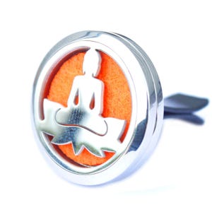 May include: A silver car air freshener featuring a Buddha silhouette on a lotus flower, set against an orange background. It has a clip for attachment. The design is circular.