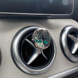 May include: A silver car air vent diffuser featuring a tree of life design. The diffuser has a teal background and is set against a silver and black car vent. The car interior has a light beige textured surface.