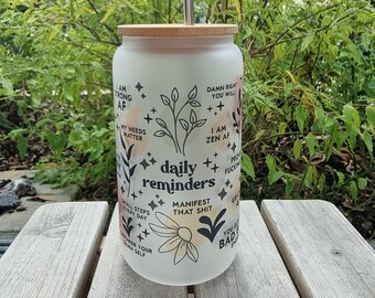 May include: White glass tumbler with a bamboo lid and a straw. The tumbler has black text and illustrations with motivational quotes like "daily reminders", "I am zen AF", "manifest that shit", and "you got this baddie".