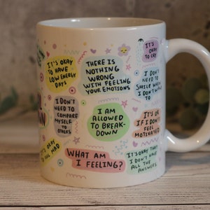 Pu&ograve; includere: Tazza in ceramica bianca con testo e illustrazioni colorate. Il testo include frasi come "Va bene avere giornate con poca energia", "Non c'&egrave; niente di sbagliato nel provare le tue emozioni", "Mi &egrave; permesso crollare" e "Cosa sto provando?"
