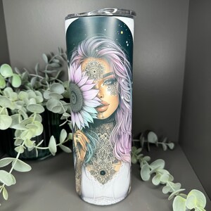 May include: A stainless steel tumbler with a black and white design featuring a woman with purple hair holding a sunflower. The woman has intricate tattoos and a mandala design on her chest.
