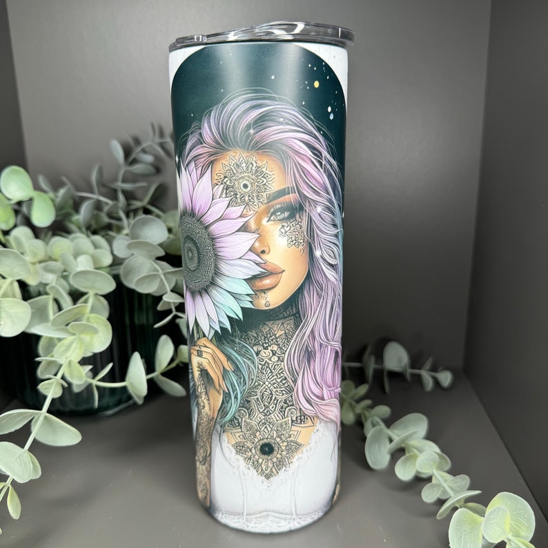 May include: A stainless steel tumbler with a black and white design featuring a woman with purple hair holding a sunflower. The woman has intricate tattoos and a mandala design on her chest.