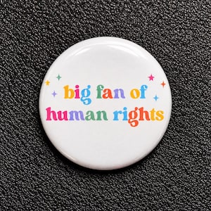 Könnte beinhalten: Ein runder, weißer Ansteckbutton mit dem mehrfarbigen Schriftzug 'big fan of human rights' in einer wellenförmigen Retro-Schriftart. Das Design wird durch kleine, bunte vierzackige Sternsymbole ergänzt, die um den Text verteilt sind.