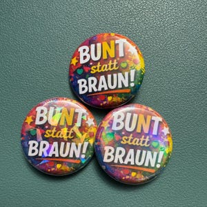 Könnte beinhalten: Drei bunte runde Buttons mit dem weißen Text "BUNT statt BRAUN!". Die Buttons haben einen lebendigen, mehrfarbigen Hintergrund mit Sternen und Herzen. Sie liegen auf einer strukturierten, dunkelgrünen Oberfläche.
