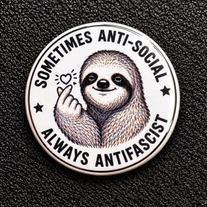 Könnte beinhalten: Ein runder Ansteckbutton mit weißem Hintergrund und einer zentralen Illustration eines braunen Faultiers, das ein Fingerherz formt. Der Text "SOMETIMES ANTI-SOCIAL" steht oben und "ALWAYS ANTIFASCIST" unten in schwarzer Schrift.