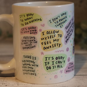 Pu&ograve; includere: Tazza in ceramica bianca con testo e illustrazioni colorate. Il testo include frasi come "Va bene voler non fare niente", "Mi permetto di sentire la mia ansia" e "Non c'&egrave; niente di male nel provare emozioni negative".