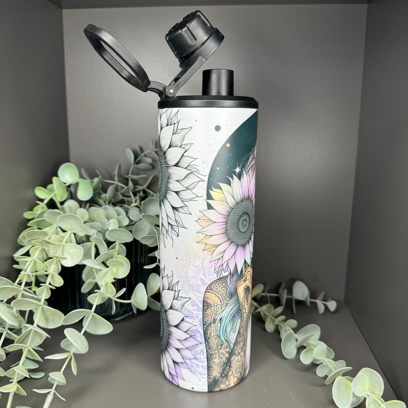May include: A white stainless steel water bottle with a black lid and a floral design featuring sunflowers and a woman's face. The design is in shades of black, white, pink, and purple.