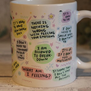 Pu&ograve; includere: Tazza in ceramica bianca con testo e illustrazioni colorate. Il testo include frasi come "Va bene avere giornate con poca energia", "Non c'&egrave; niente di sbagliato nel provare le tue emozioni", "Mi &egrave; permesso crollare" e "Cosa sto provando?"